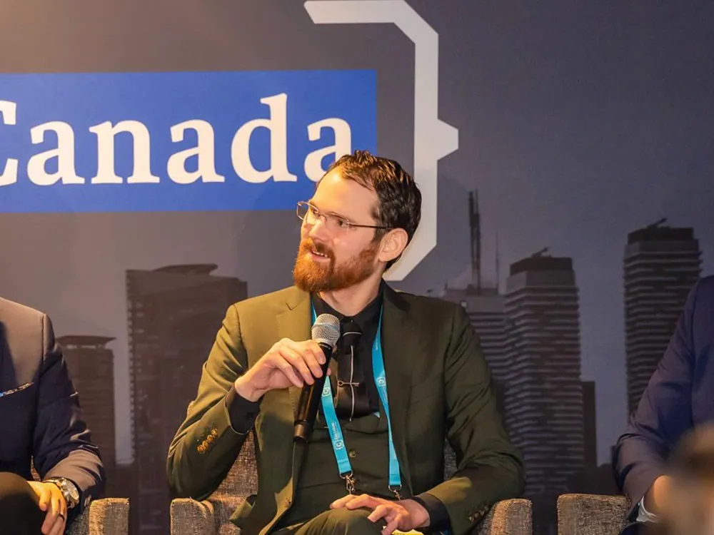 Speaking on a panel at a conference, seated with a microphone in front of a backdrop showing a city skyline.